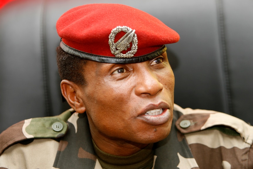 A middle-aged African man in a military uniform with a red beret speaks while seated in a leather chair.