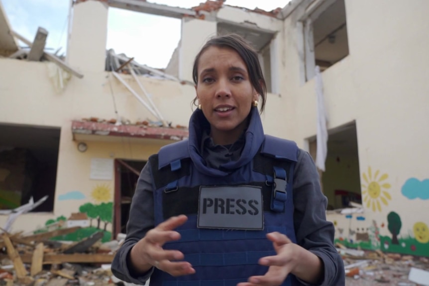 Woman wearing bullet-proof vest standing in ruins of a building.