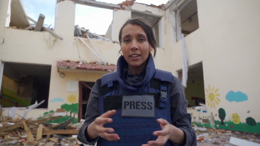 Woman wearing bullet-proof vest standing in ruins of a building.