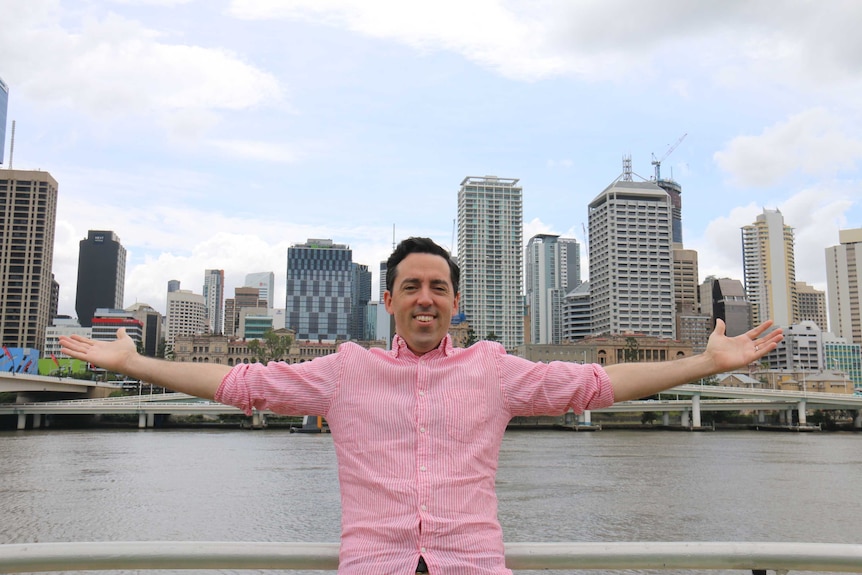 Comedian Mark Lombard poses with his arms extended in front of Brisbane CBD and river