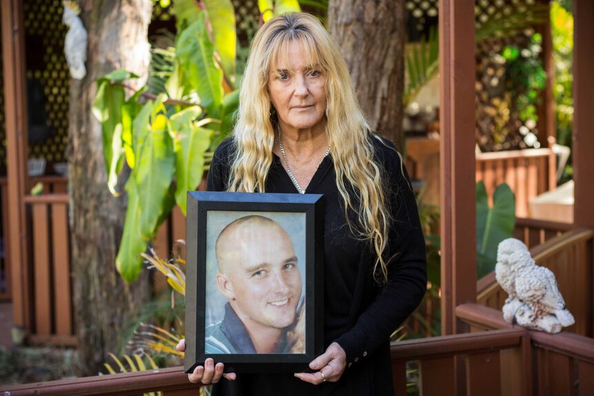 Blonde woman stands outside on deck holding photo of shaved head young man.