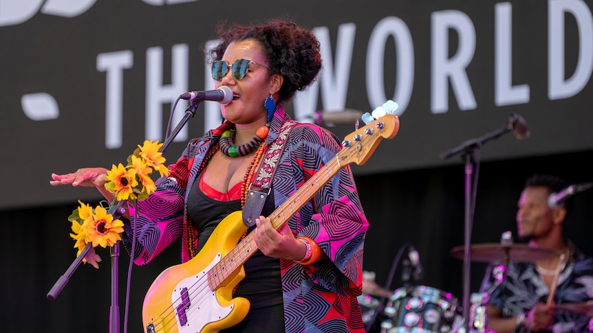 A woman wearing bright colours holds an electric bass while singing into a microphone.
