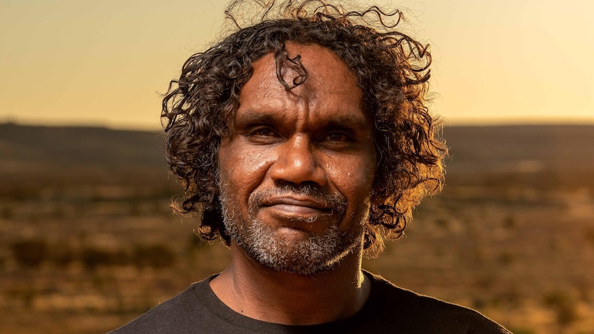 A dark-skinned man with curly hair looks confidently into the camera, with a desert landscape behind him.