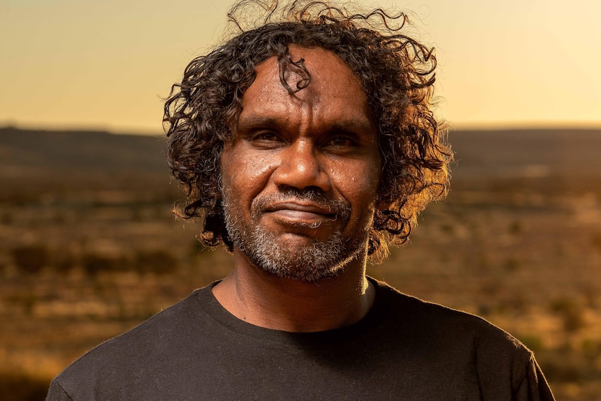 A dark-skinned man with curly hair looks confidently into the camera, with a desert landscape behind him.
