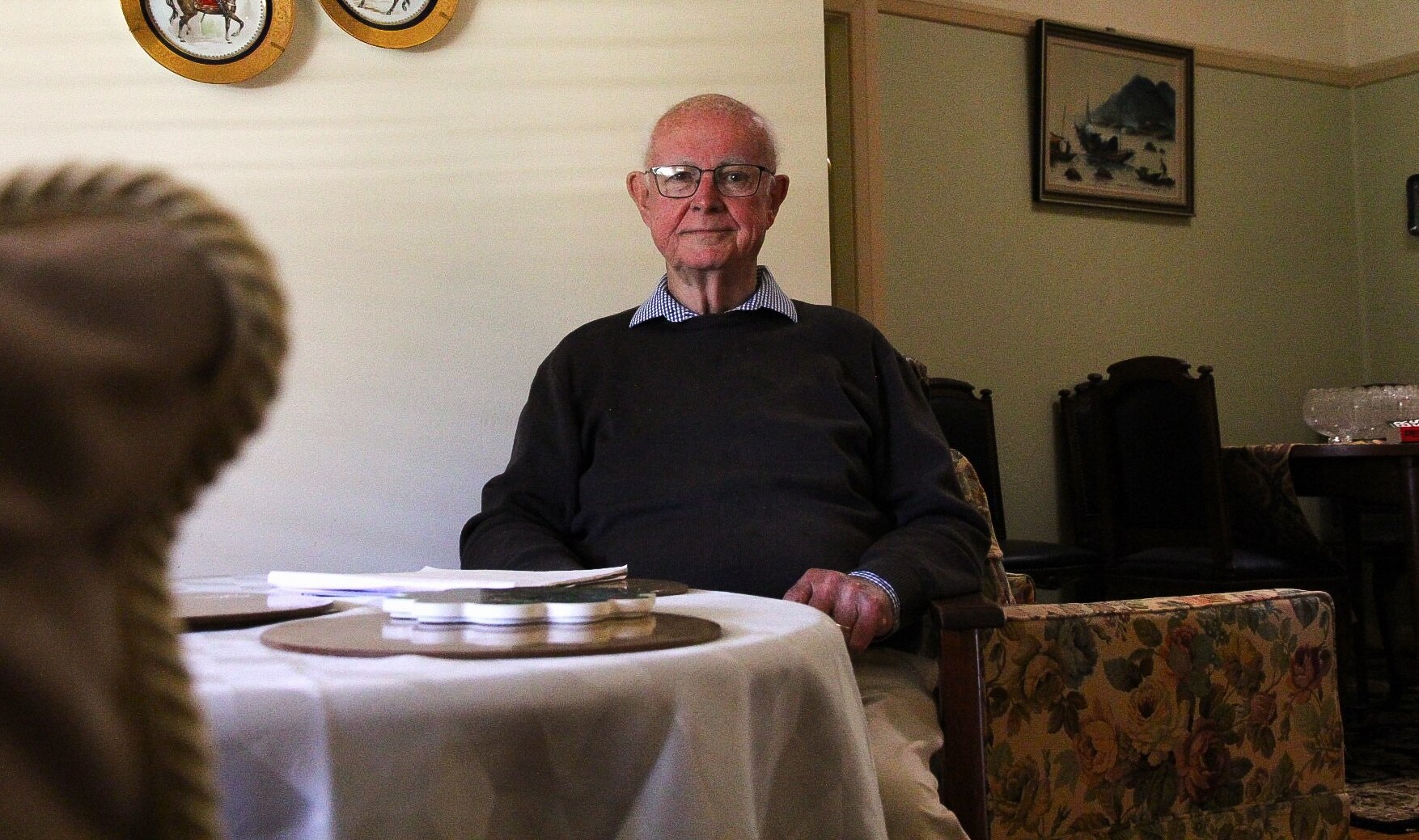 An elderly man sitting behind a table with a vintage chair in the foreground. 