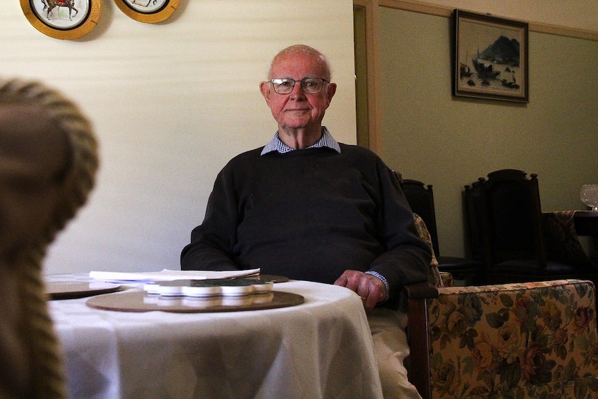 An elderly man sitting behind a table with a vintage chair in the foreground. 