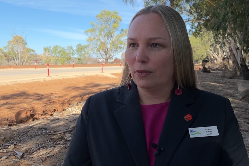 A woman with blonde hair wears a pink top with a black blazer, she wears red poppy. She looks solemnly with roadworks behind her