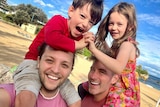 Sean Szeps and husband Josh Szeps smiling on a sunny beach with twin kids on their shoulders