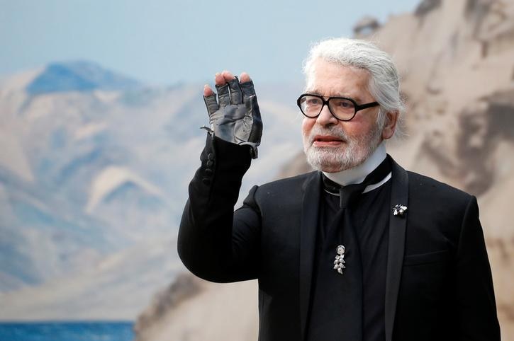 Karl Lagerfeld wears a black suit, glove and glasses. He waves to the crowd with a mountain backdrop behind him.