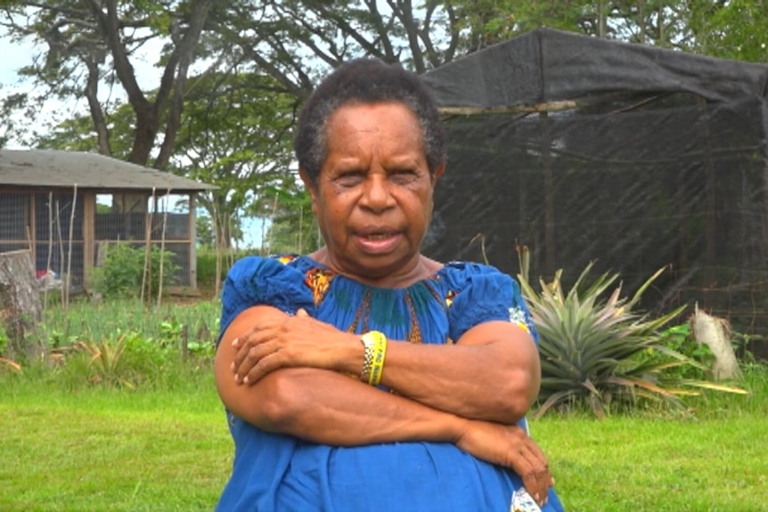 Woman in blue dress talks to camera, sitting in open green field surrounded by trees, plants and garden structures.  