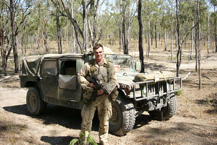 Dr Steven Scally, in uniform, standing in front of an army vehicle and holding a weapon.