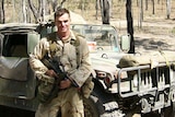 Dr Steven Scally, in uniform, standing in front of an army vehicle and holding a weapon.