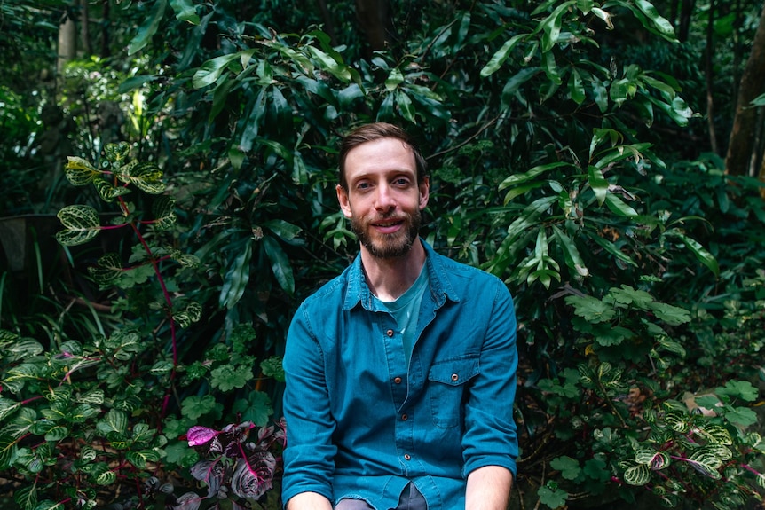 A man in a blue button up shirt sitting in front of lush green foliage