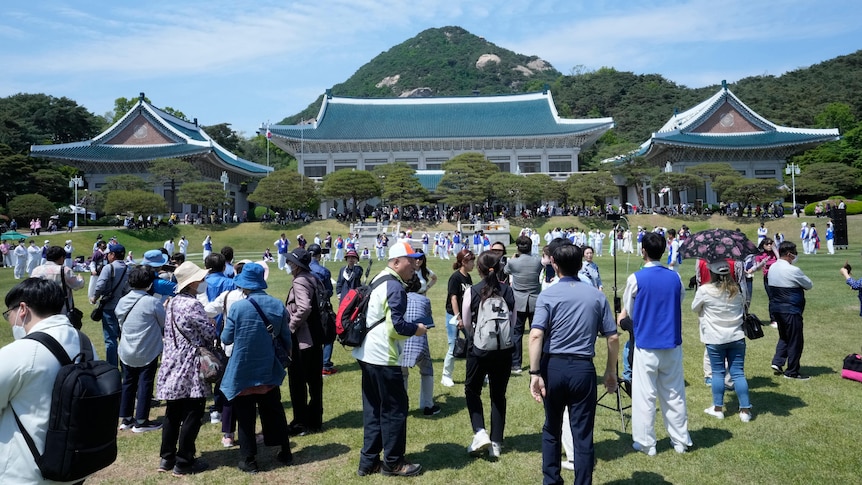 A large crowd gathers on the large front lawn of an Oriental-looking building with a blue roof