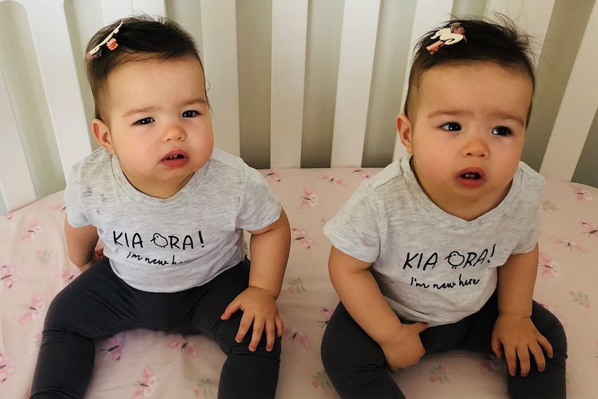 Two identical twin girls with brown hair sitting in a white cot wearing grey t shirts and with seahorse bows in their hair