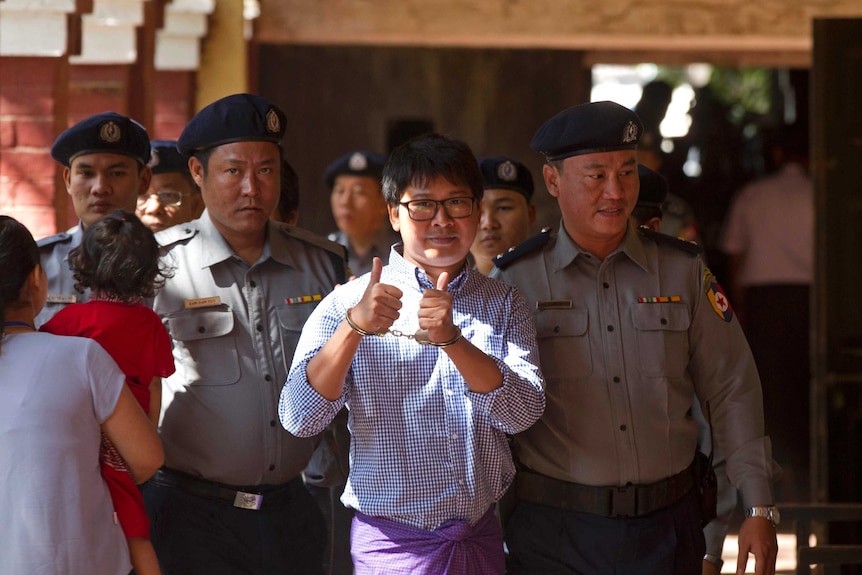 Wide shot of a man giving thumbs up as he is led by guards.