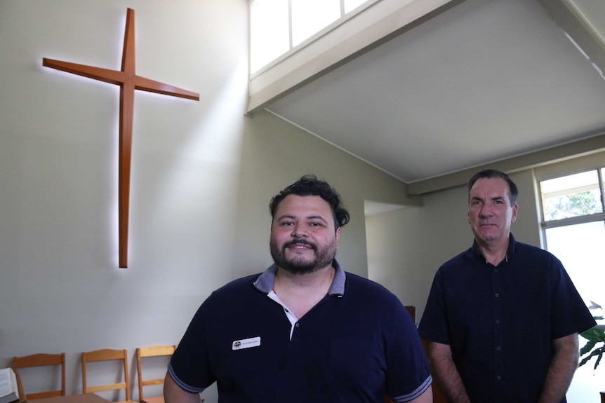 A short bearded man stands in front of taller older man inside church with cross and sunlit windows.