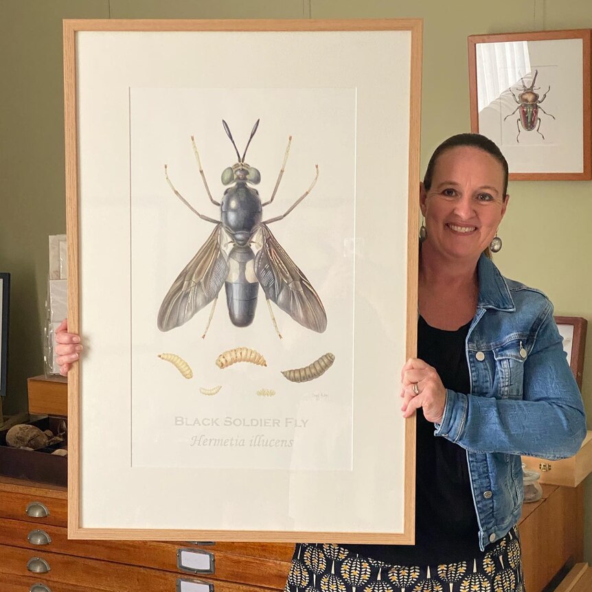 A woman smiles as she holds up a large frame containing a painting of a beetle.