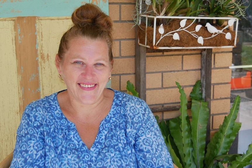 A woman sits in front of a brick wall wearing a blue top and smiles at the camera. Her  hair is tied up and she has blue eyes.