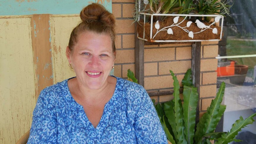 A woman sits in front of a brick wall wearing a blue top and smiles at the camera. Her  hair is tied up and she has blue eyes.