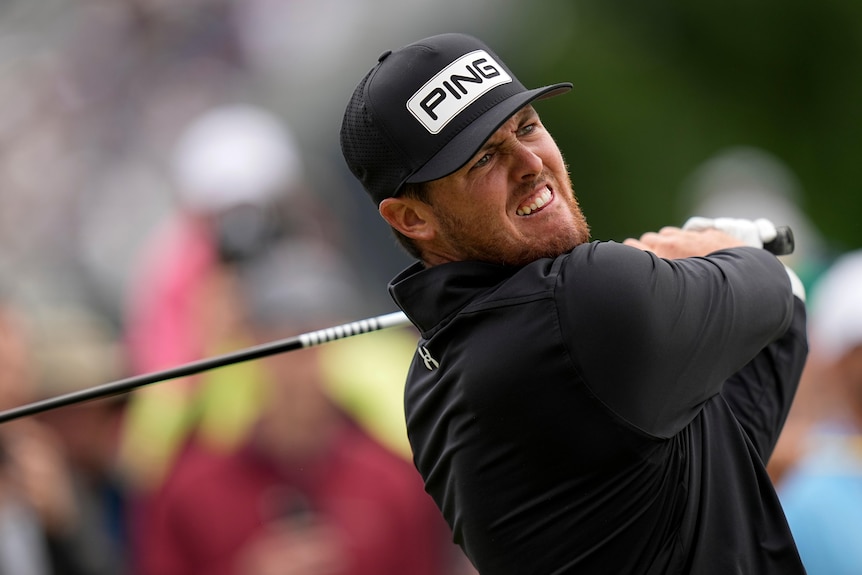 A golfer in a black cap and shirt looks down the fairway as he follows through from his tee shot.