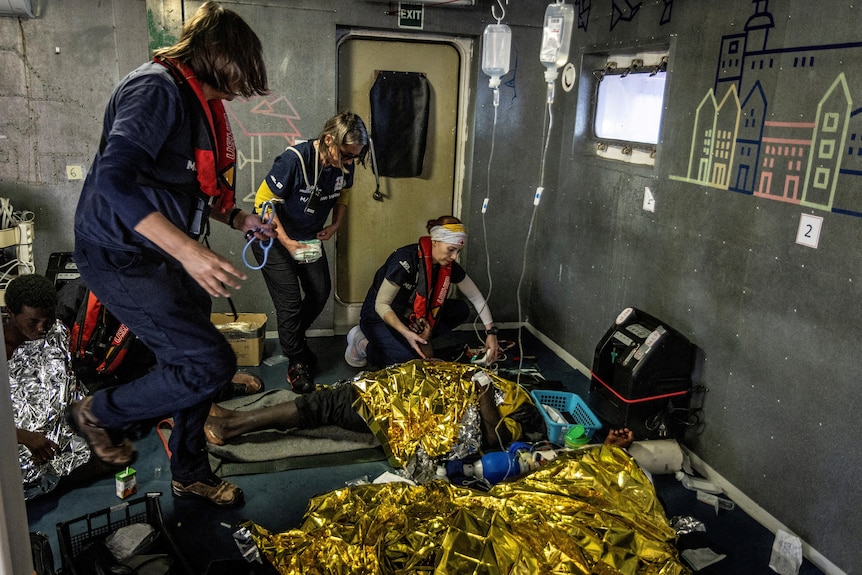 Three women provide first aid to two people who are on the floor of a ship covered in gold blankets