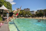 Two boys fling themselves into an inviting pool under clear a blue sky with high-rise buildings in the background