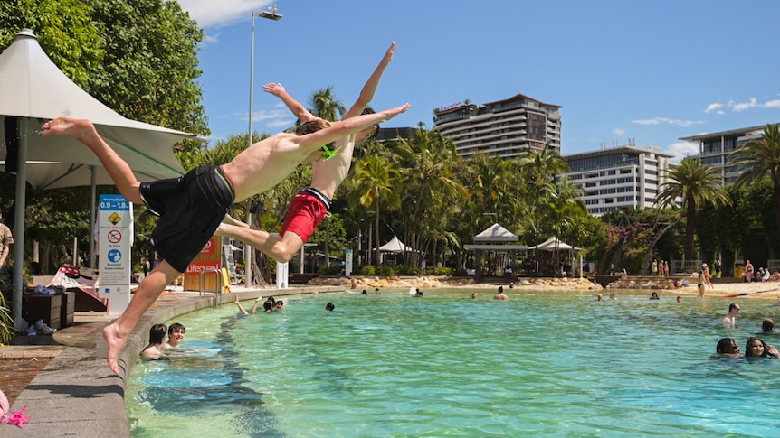 Two boys fling themselves into an inviting pool under clear a blue sky with high-rise buildings in the background