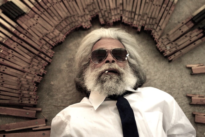 A bearded man wearing sunglasses lies on the floor surrounded by piano keys.