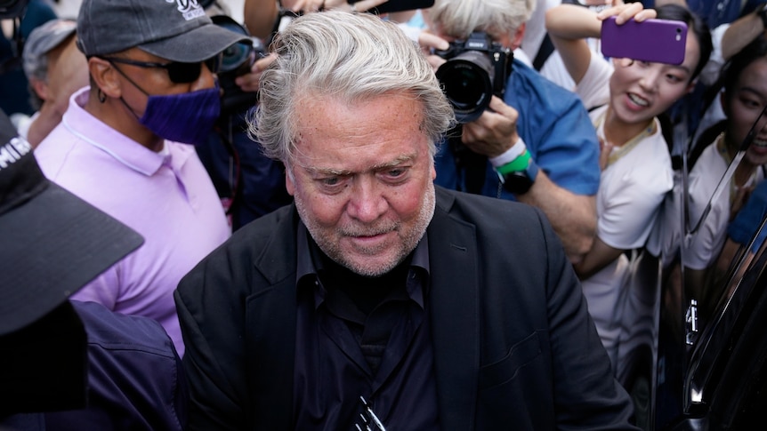 Steve Bannon is wearing a black suit while getting into a car, surrounded by a crowd with camera's and phones aimed towards him.