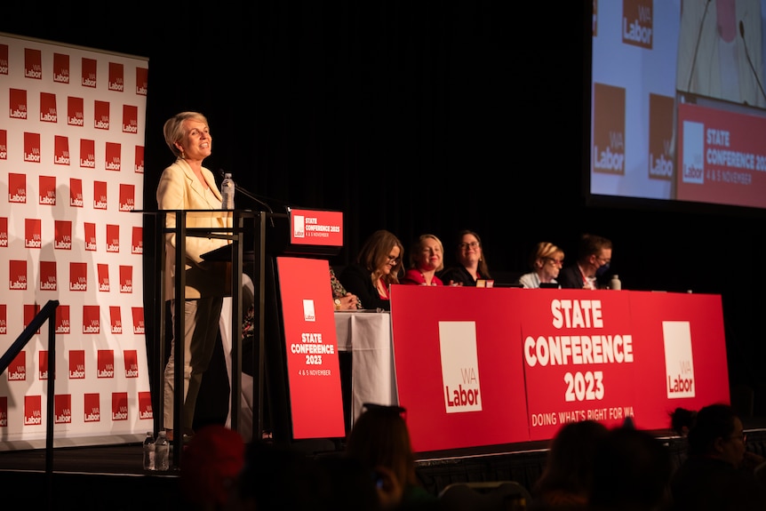 A woman with short blonde hair speaks from a lectern next to a table with a red sign in front.