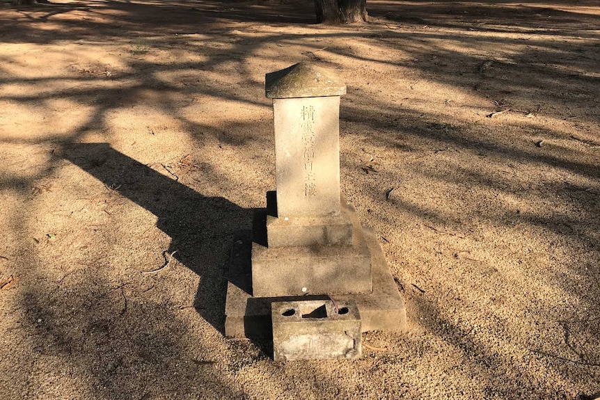 A lone headstone with Japanese script on it stands in a dry, gravel covered park.