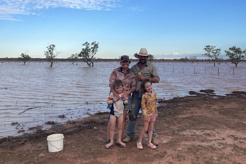 A family with two parents and two kids stand beside calm outback floodwaters on their property