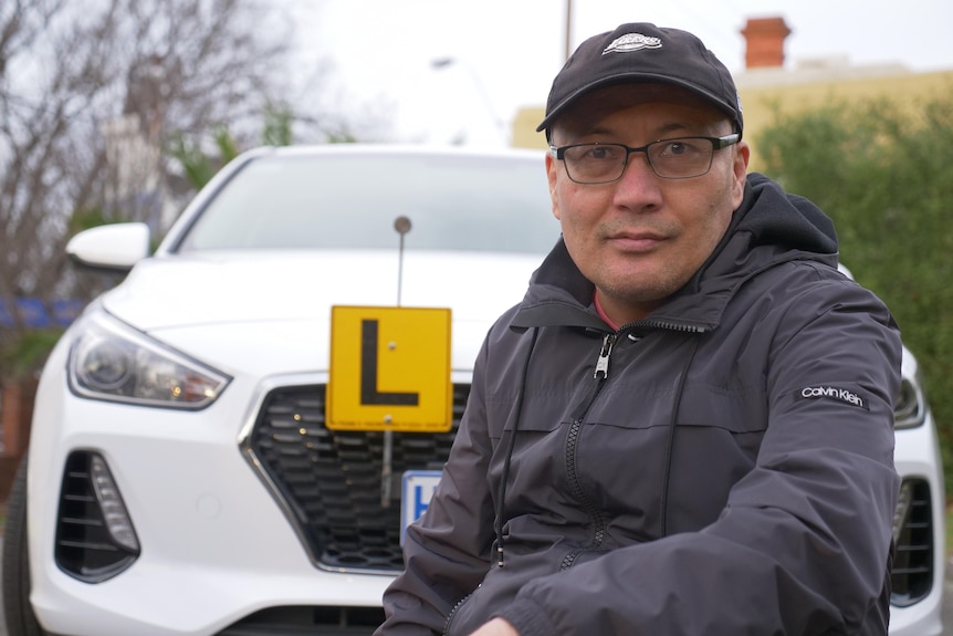 A man wearing a cap crouched in front of a car that has an L plate on it