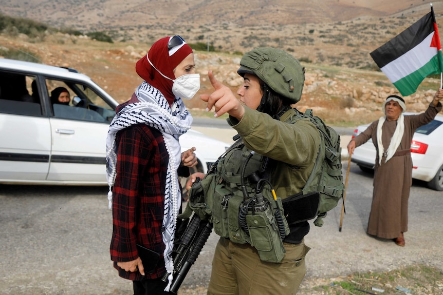 A female soldier gestures to a woman in a face mask and headscarf on a road