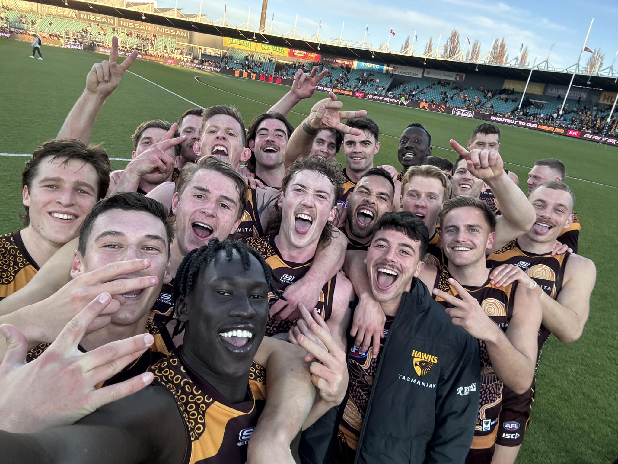 A group of footballers in jerseys huddle together for a group selfie on a footy pitch.