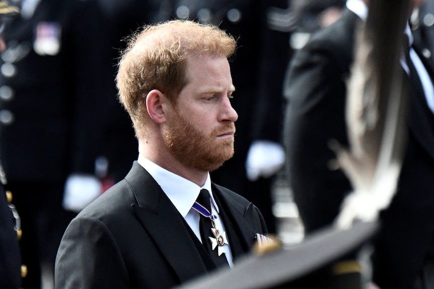 Prince Harry with a beard looks down while wearing a dark suit and tie.