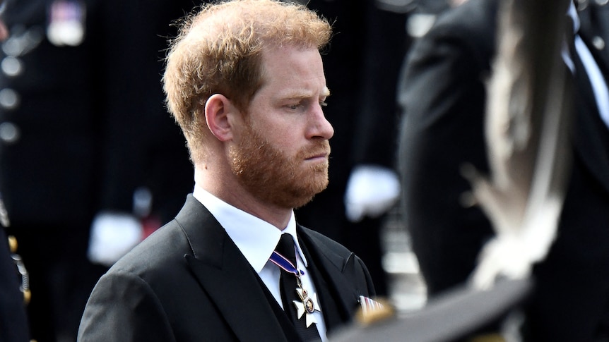 Prince Harry with a beard looks down while wearing a dark suit and tie.