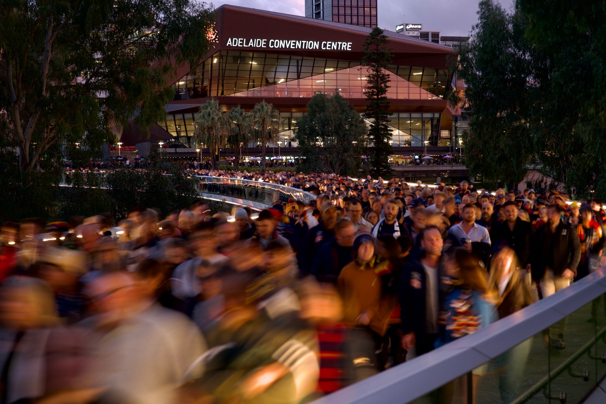 Uma longa exposição desfocada de uma multidão de pessoas em camisetas esportivas caminhando ao longo de uma ponte metálica curva com eucaliptos