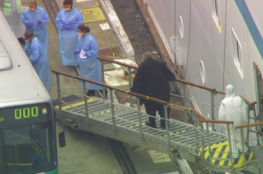 A man wearing a black top walks down a gangway from a cruise ship to a bus