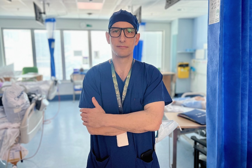 Doctor in scrubs standing in a ward with arms crossed and not smiling