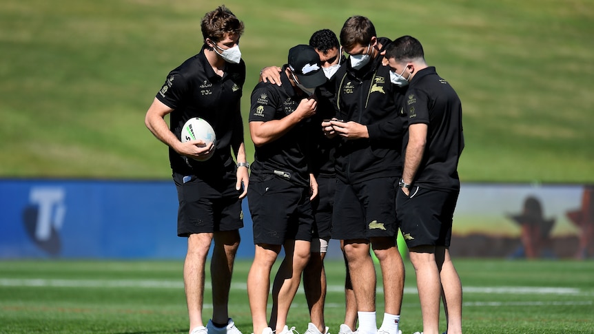 A group of rugby players in black wearing face masks hold the ball in middle of grassy field.