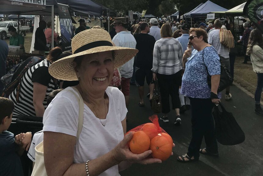 A woman stands in front of other marketgoers, smiling, wearing a hat, and holding oranges.