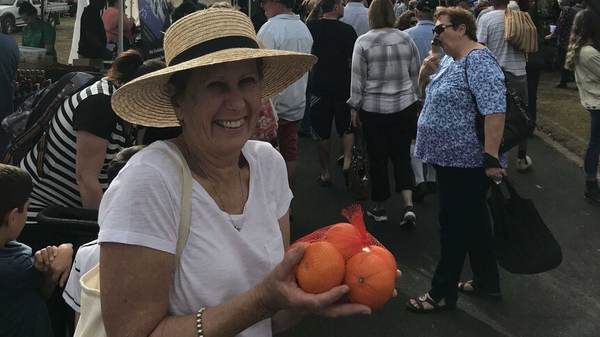A woman stands in front of other marketgoers, smiling, wearing a hat, and holding oranges.