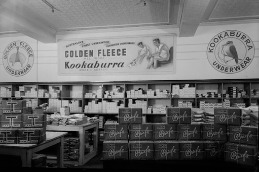 A black and white image of a shop with Golden Fleece ads on the walls above shelves with boxes of woollen underwear