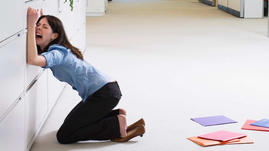 A businesswoman in a blue shirt leaning on a filing cabinet while crying.