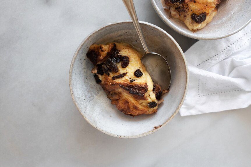 A serving of bread and butter pudding in a ceramic bowl  with a spoon, a second bowl in the background. A comforting dessert.