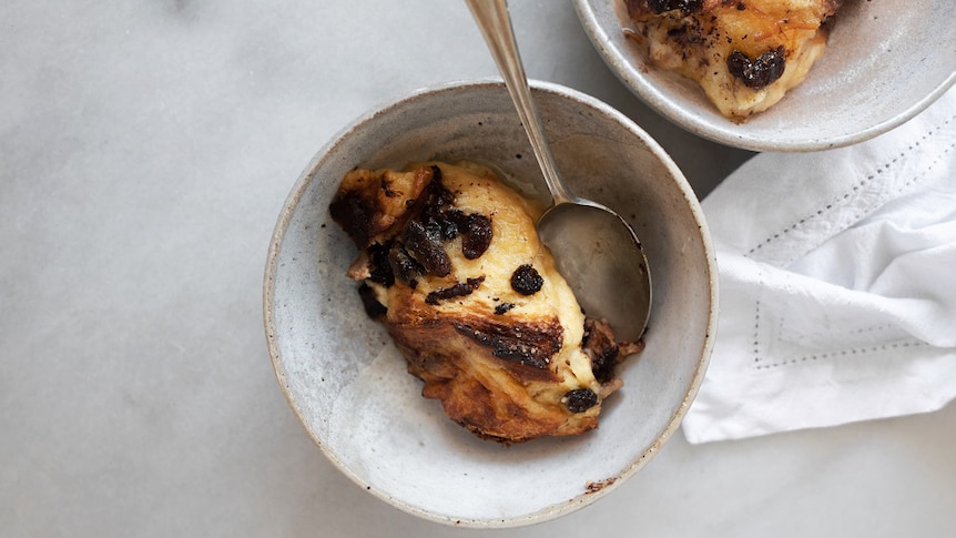 A serving of bread and butter pudding in a ceramic bowl  with a spoon, a second bowl in the background. A comforting dessert.