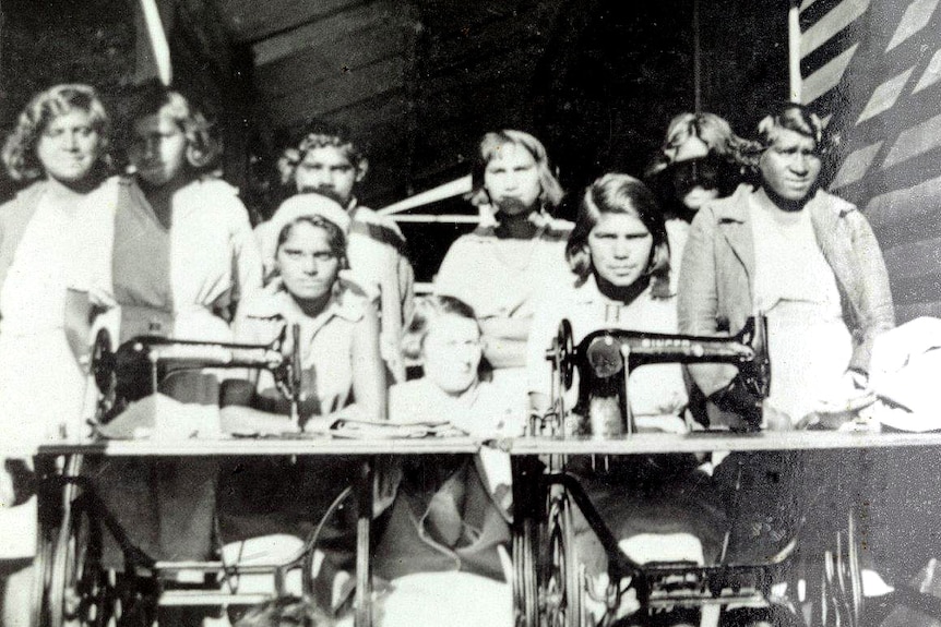 A group of children stand behind two girls sat at old Singer sewing machine tables.