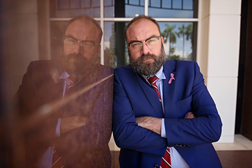 A bearded balding man in blue suite, red tie, wears glasses, leans against a highly reflective building, arms crossed, serious.